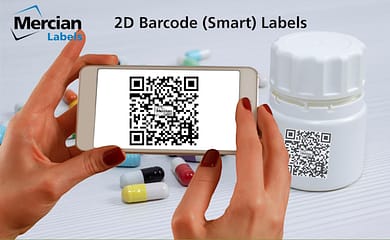 Two hands holding a smartphone with the image of a 2D (QR Code style) barcode on the phone. In the background is a pill bottle with the same 2D QR code style barcode on it and a few coloured pill capsules spread out on the table to the side of it.